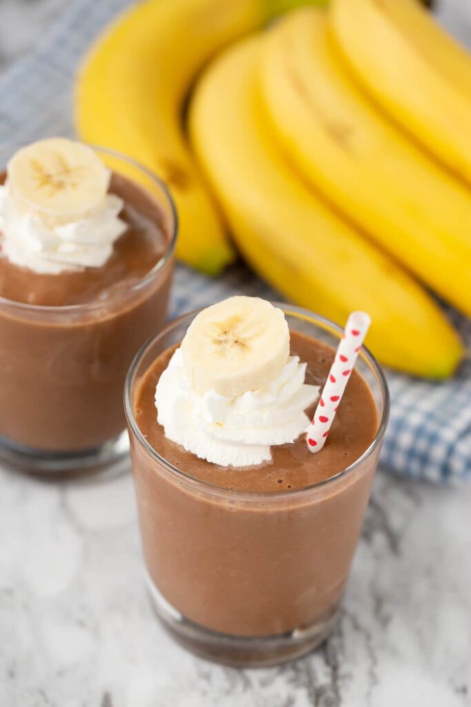 Chocolate Banana Smoothie in two glasses with a straw in it with whip cream on top.