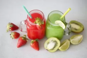 Two Smoothies one red one green in glasses side by side with straws surrounded by strawberries and kiwis