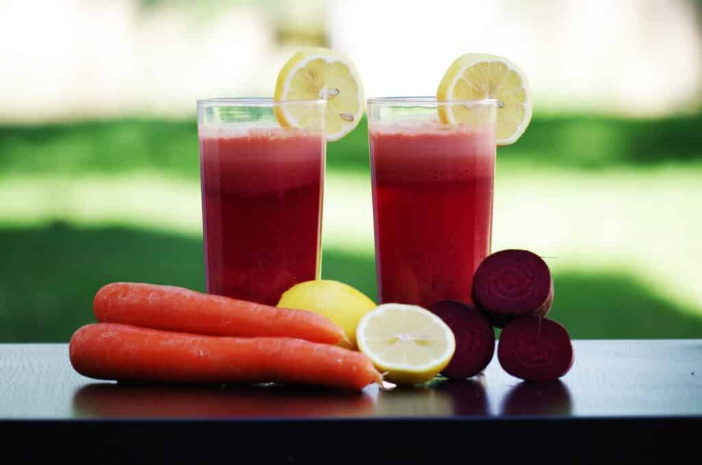 Two red twenty one day smoothies sitting on a table with carrots, fruit and lemons.