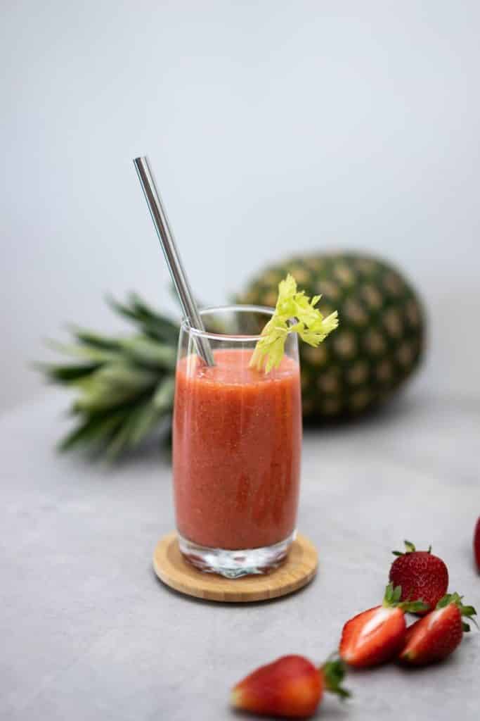 A strawberry smoothie with a straw in it and a whole pineapple in the background.