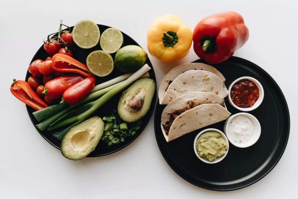 Healthy fats are avocados, tacos made with grains, yellow and red peppers, spring onions, and limes displayed on round black plates.