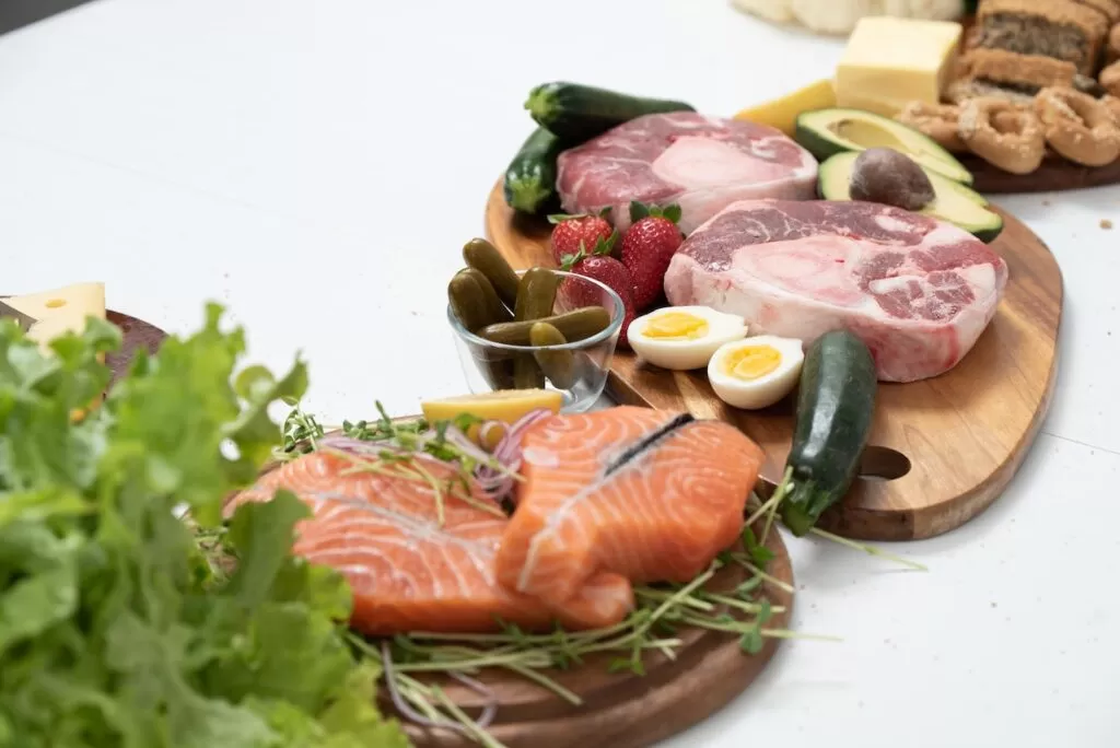 Major food groups salmon, beef steaks, sliced boiled eggs, green lettuce, cucumber, and strawberries displayed on round wooden plates.