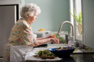 Elderly blonde haired woman washing vegetables under running water from a kitchen faucet.