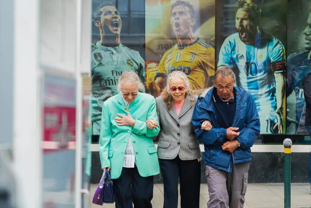 Three elderly people two women and a man walking arm in arm in front of human posters.