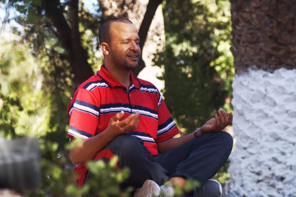 A bearded man wearing a red and white shirt meditating outside.