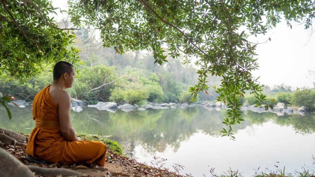 A Monk practicing yoga and meditation outdoors by a lake under a tree.