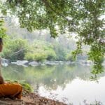 A Monk practicing yoga and meditation outdoors by a lake under a tree.