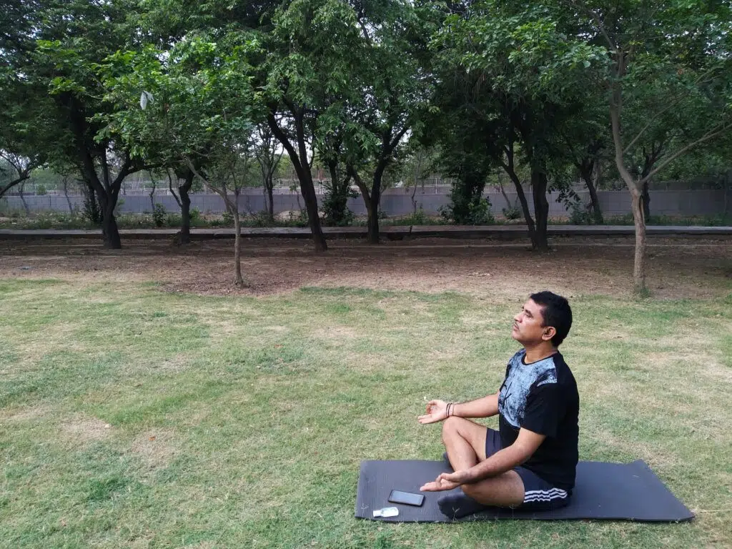 A man sitting with his leg crossed in a yoga position on a yoga mat outside meditating.