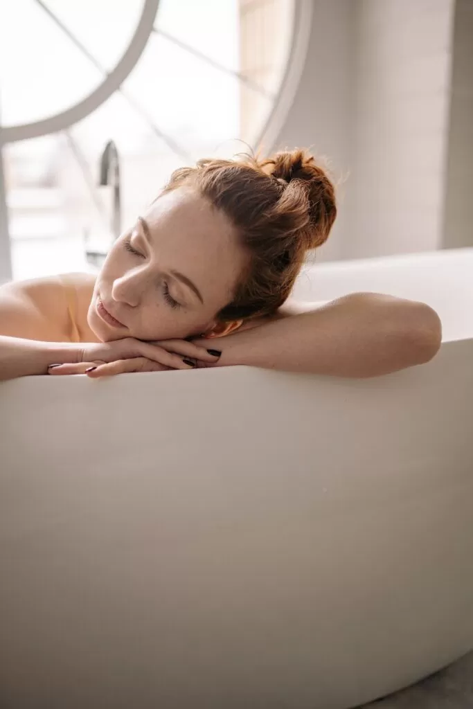 A woman sleeping with her head on her hands on the side of the tub.