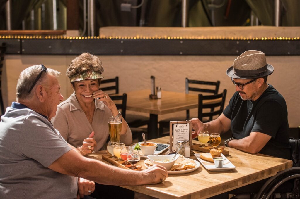 three smiling people sitting at a table with plate of foods and drinks having social interaction with mealsl