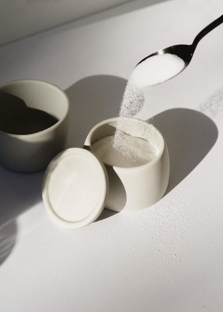 A spoon of white sugar poured into a white ceramic mugs on white table